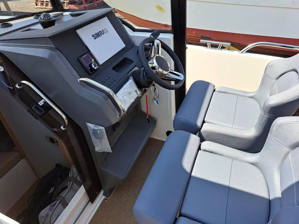Boat cockpit featuring a steering wheel, SIMRAD display, control panel, and two gray seating areas, with a spacious design.